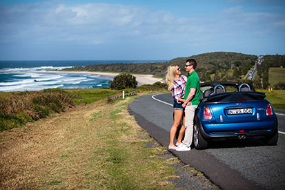 Couple leaning on car along Coast Road with beach in background