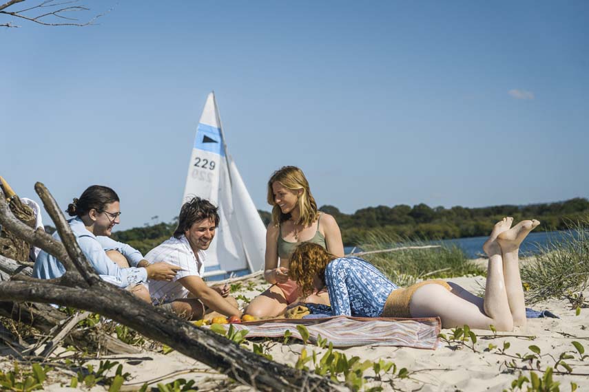 Island Life 2 - Couples lying on sand playing cards with yacht in background