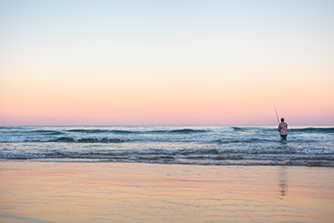 Pastel coloured sunset with man fishing in water at beach