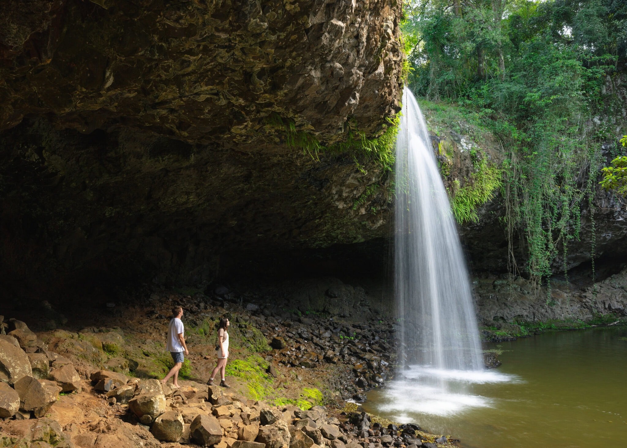 Killen Falls, Ballina Hinterland – Rainforest Escape with a Hidden Waterfall Two people standing beneath the dramatic overhang of Killen Falls in the Ballina hinterland, surrounded by rainforest and a flowing waterfall.