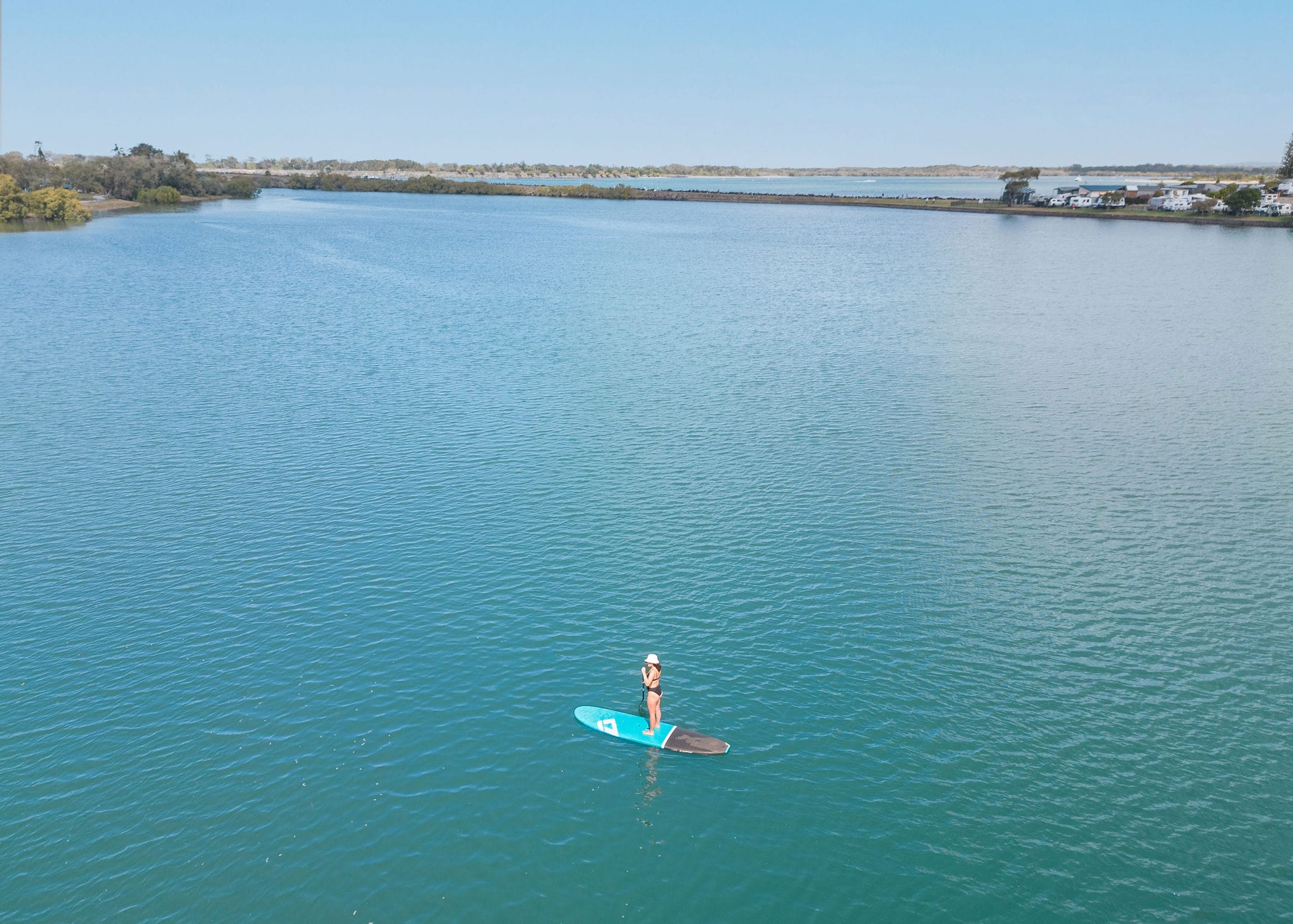Stand-Up Paddleboarding Serenity at Shaws Bay, Ballina A woman on a stand-up paddleboard glides across the calm, turquoise waters of Shaws Bay in Ballina under a clear blue sky.