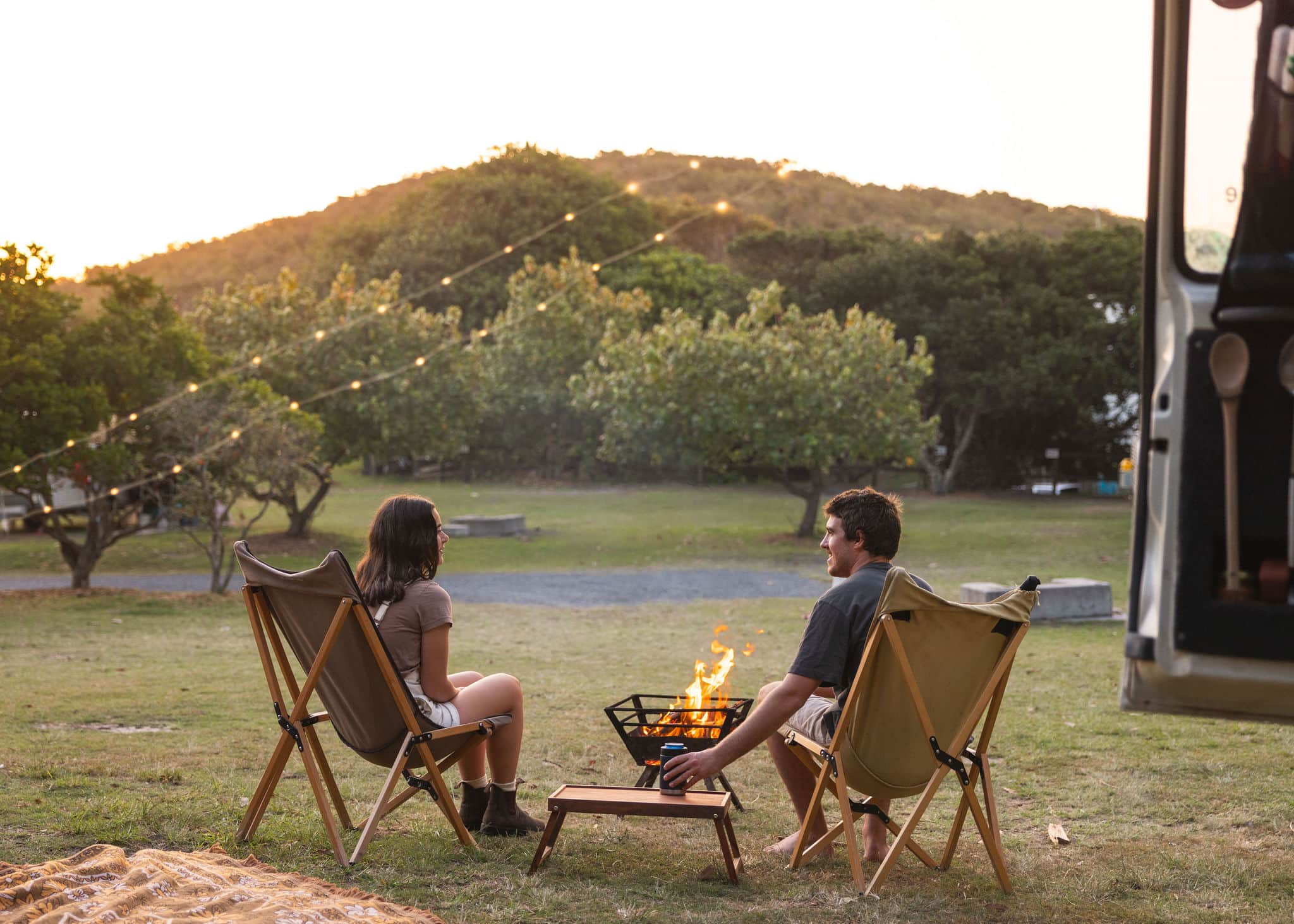 Campfire Sunsets at Flat Rock Tent Park – Ballina Beachside Bliss Couple sitting by a campfire under string lights at Flat Rock Tent Park in Ballina, enjoying a peaceful sunset surrounded by trees and hills.