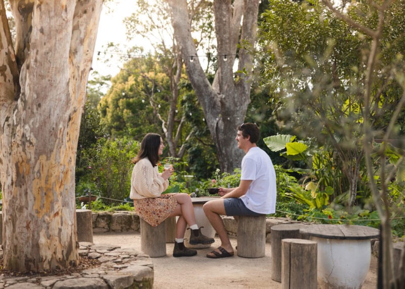 A couple enjoying coffee in a lush garden setting at a Newrybar café in the Byron Bay hinterland, surrounded by native trees and greenery.