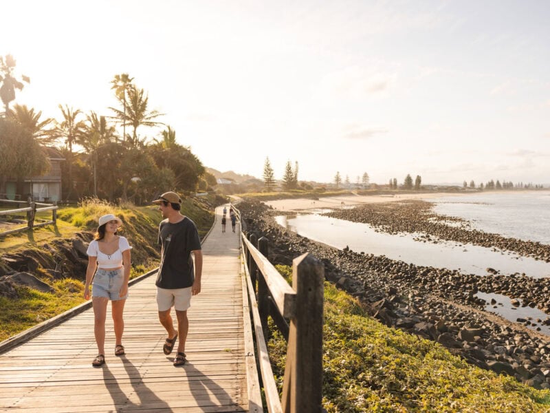 Couple walking along the coastal boardwalk at Lennox Head at sunset, with ocean views, rocky shoreline, and palm trees in the background.