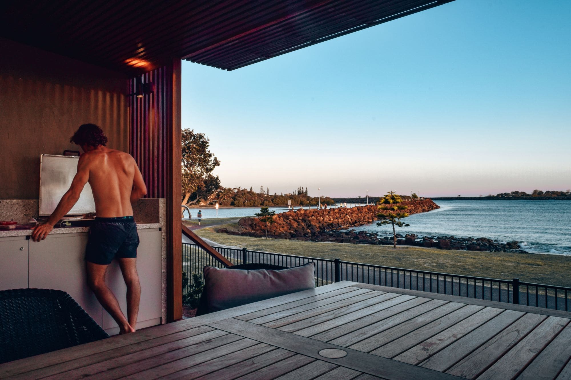 BBQ with a View – Reflections Holiday Park Shaws Bay, Ballina NSW Man cooking at a BBQ in a waterfront cabin at Reflections Holiday Park Shaws Bay, Ballina, with views over the Richmond River at sunset.