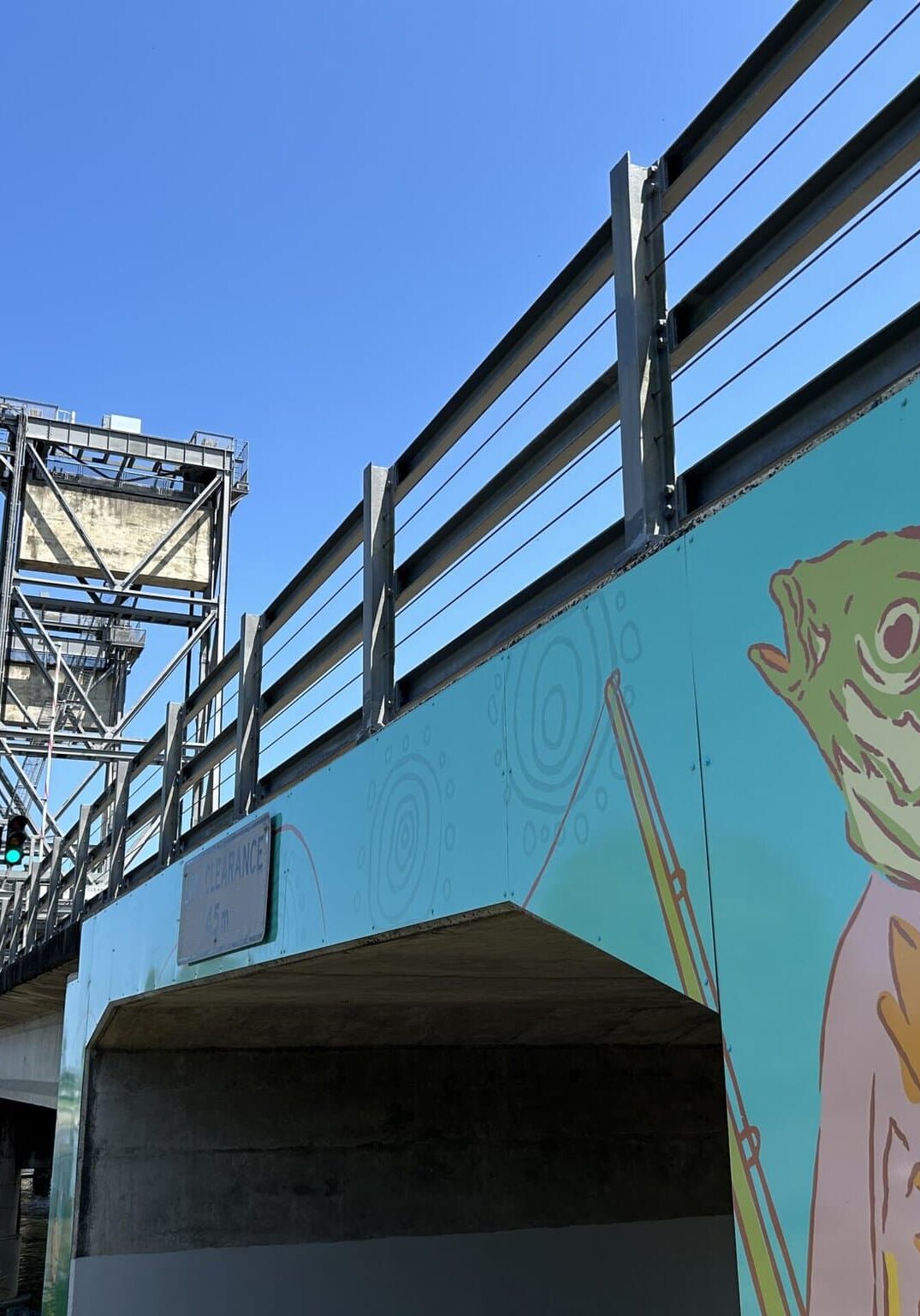 Wardell Bridge Mural: Where Art Meets the River Public mural of a fish-headed figure in a suit beneath the Wardell lift bridge, with bright blue sky and river in view.