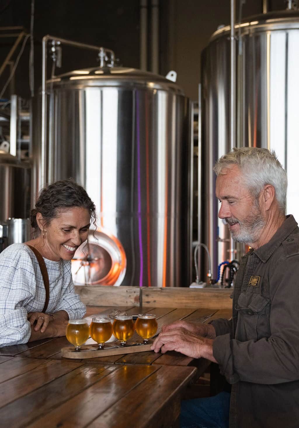 Two people enjoying a beer tasting paddle at a brewery in Ballina, with stainless steel brewing tanks in the background.