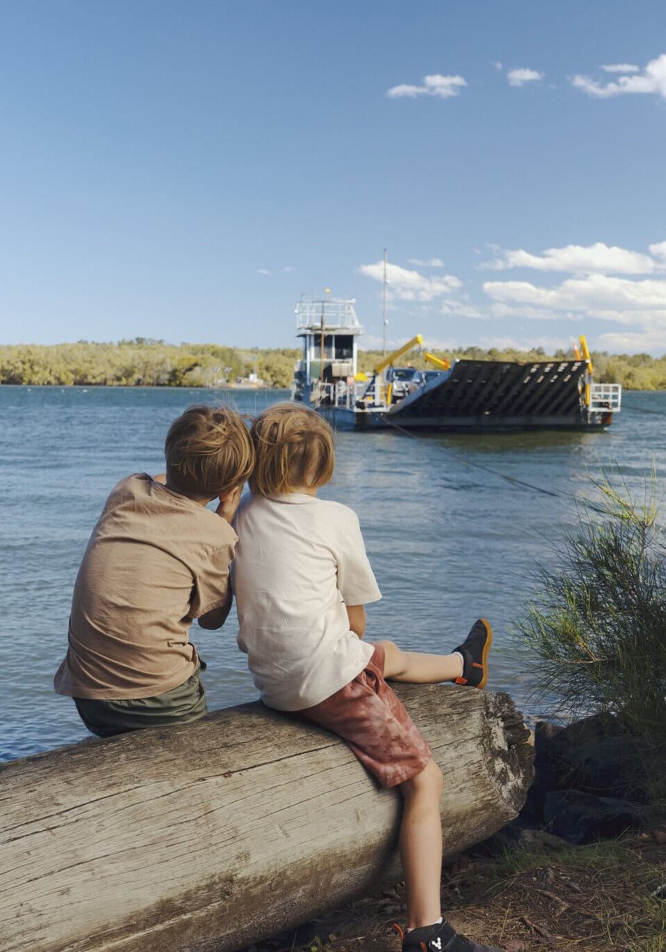Nostalgic Moments at the Burns Point Ferry, Ballina Two children sit on a log by the riverbank, watching the Burns Point Ferry cross the Richmond River in Ballina on a sunny day.
