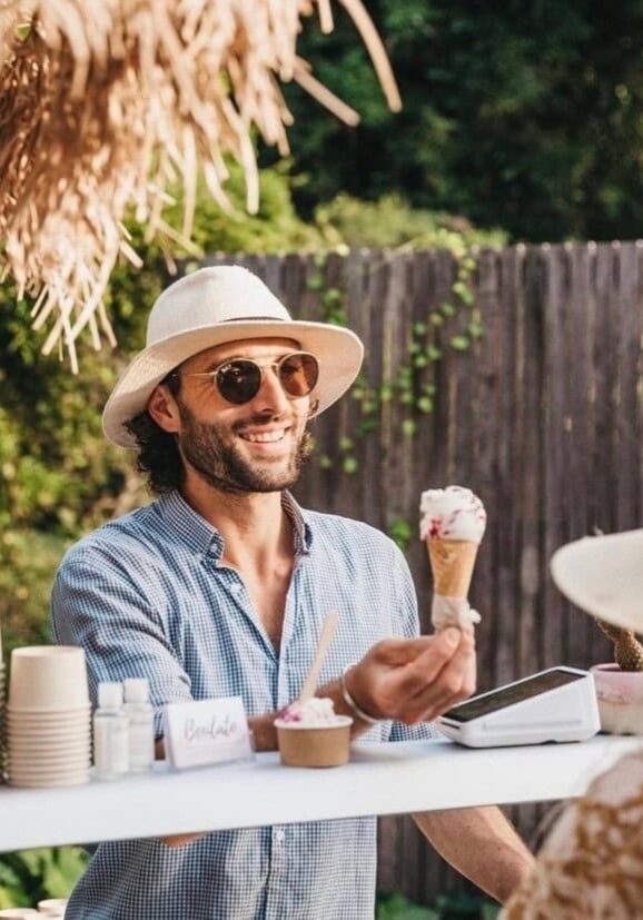 A smiling vendor at Benilato serves handcrafted gelato in a cone to a marketgoer at the Newrybar Twilight Market, surrounded by rustic décor and natural shade.