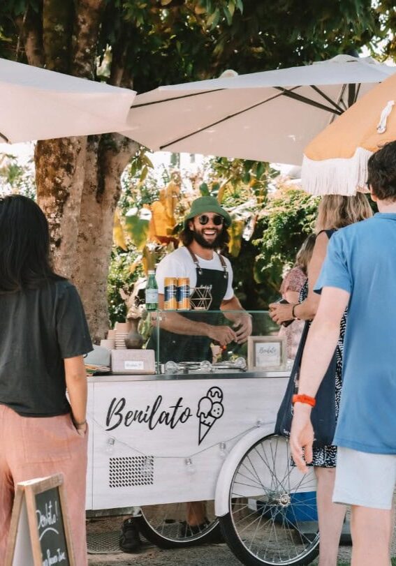 Benilato Cart at Newrybar Twilight Markets Smiling vendor serving gelato from a Benilato cart under umbrellas at the Newrybar Twilight Markets, surrounded by customers.