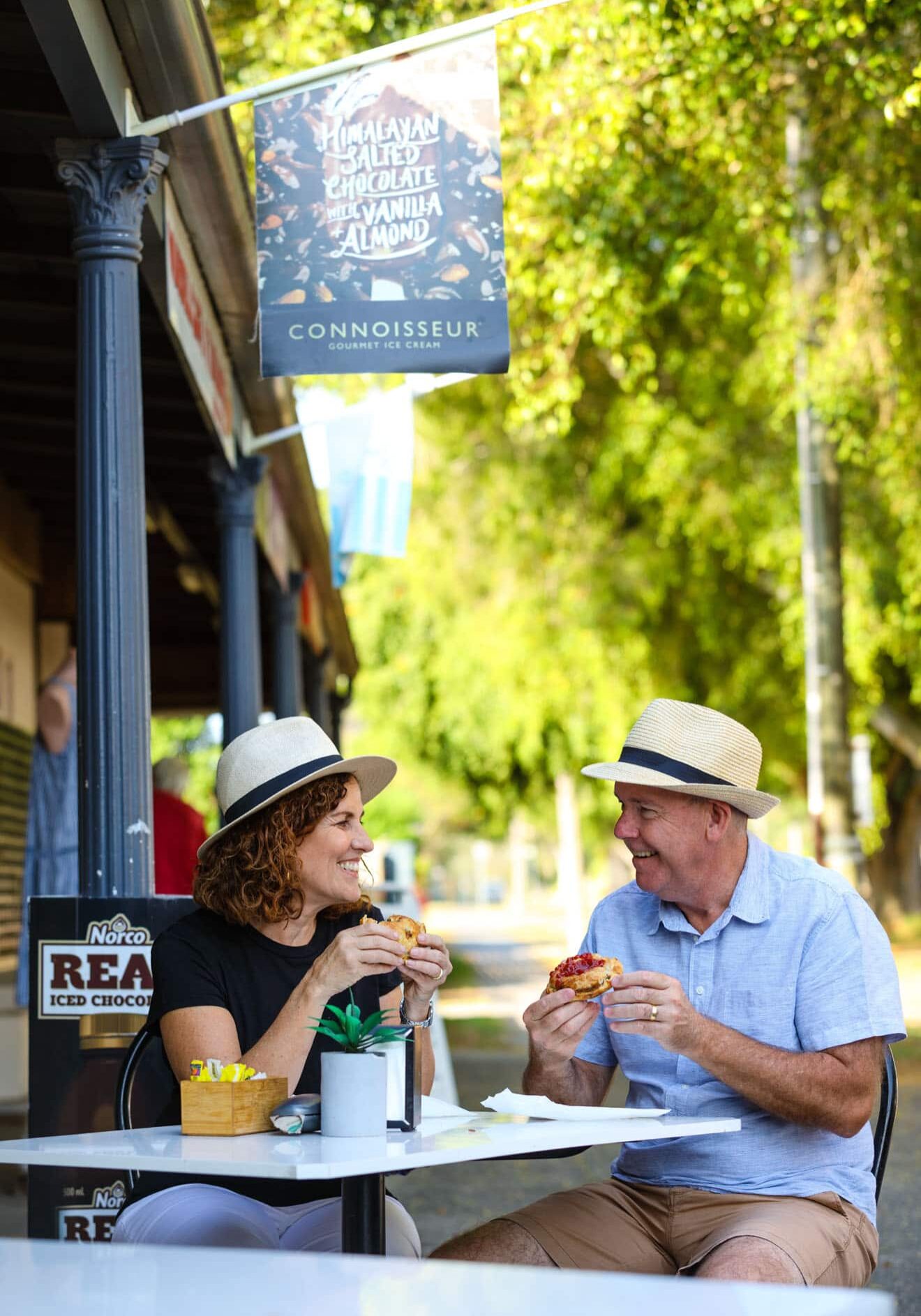 Tucking Into Wardell’s Famous Pies Couple smiling and enjoying freshly baked pies at an outdoor table in Wardell, surrounded by leafy streets and heritage shopfronts.
