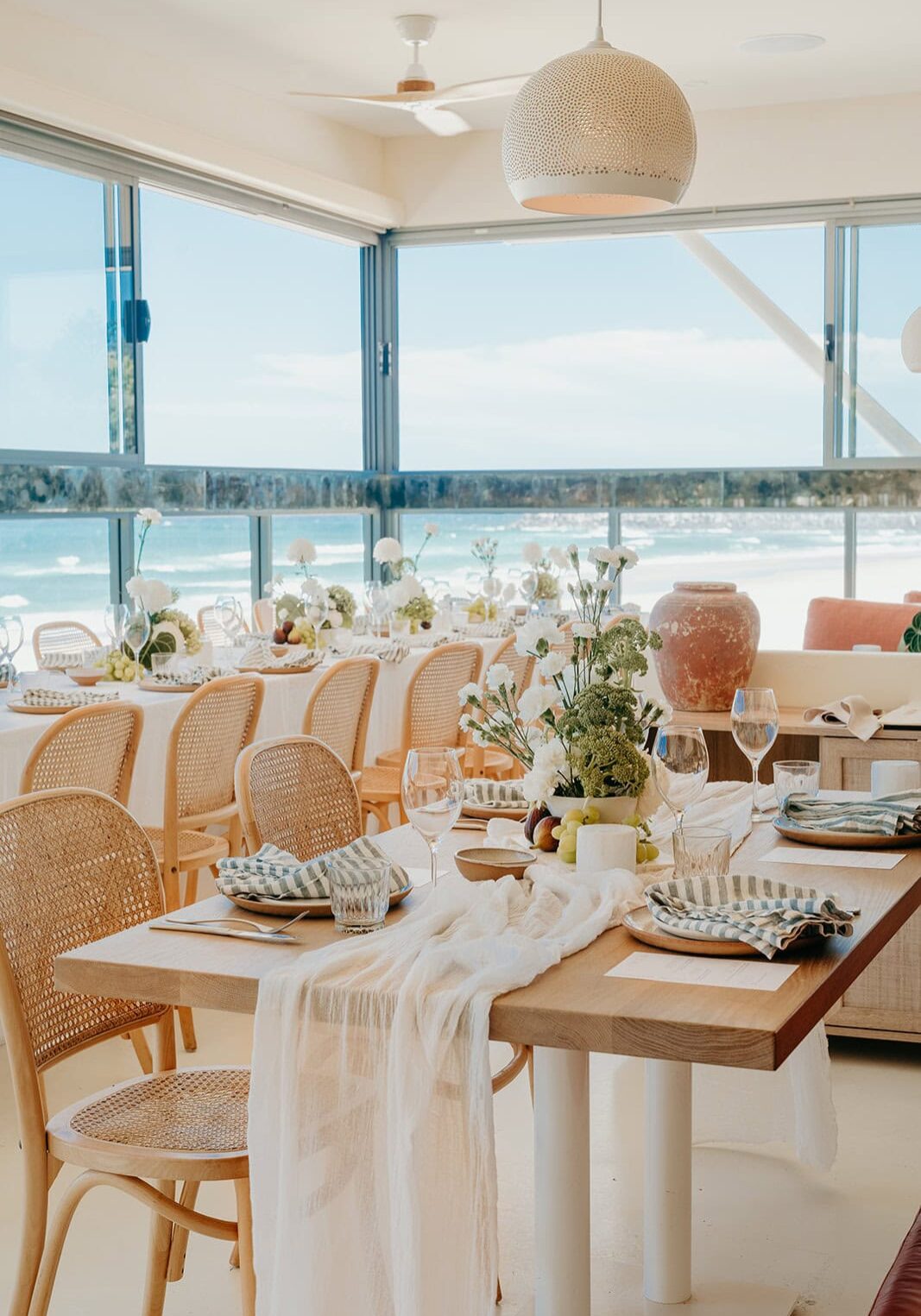 Coastal dining setup inside Capiche in East Ballina, with long tables, rattan chairs and ocean views. Title