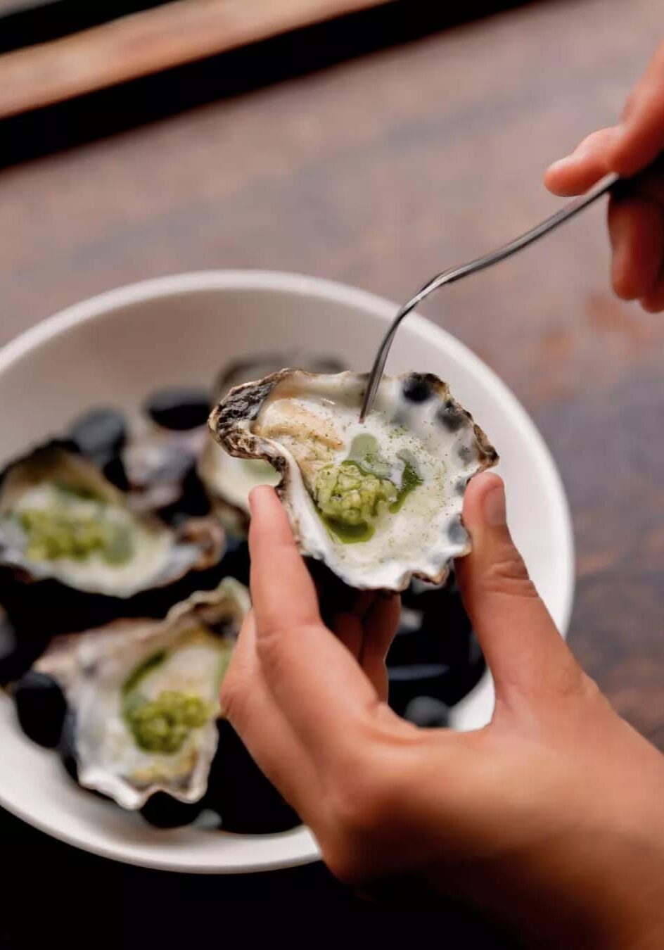 Coastal Flavours, Elevated – Dining at Shelter Lennox Head Close-up of a hand holding a freshly shucked oyster topped with green garnish at Shelter restaurant in Lennox Head.