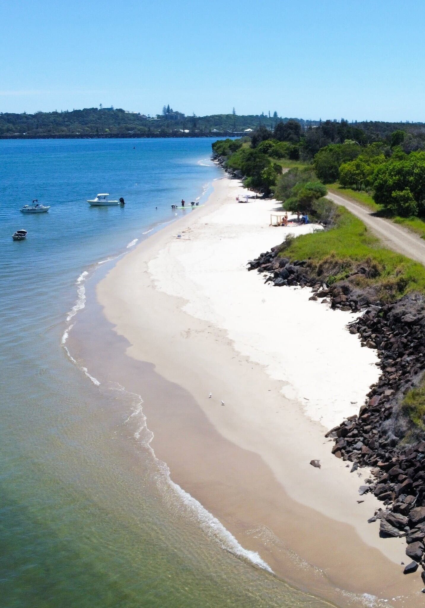 Mobbs Bay Beachfront – South Ballina, NSW Aerial view of Mobbs Bay in South Ballina, featuring calm turquoise waters, anchored boats, a sandy shoreline, and a coastal track bordered by native bushland.